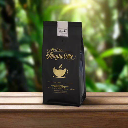 A black package of Amyta Coffee Smooth Arabica with a gold and white label, placed on a wooden surface with a lush green background.