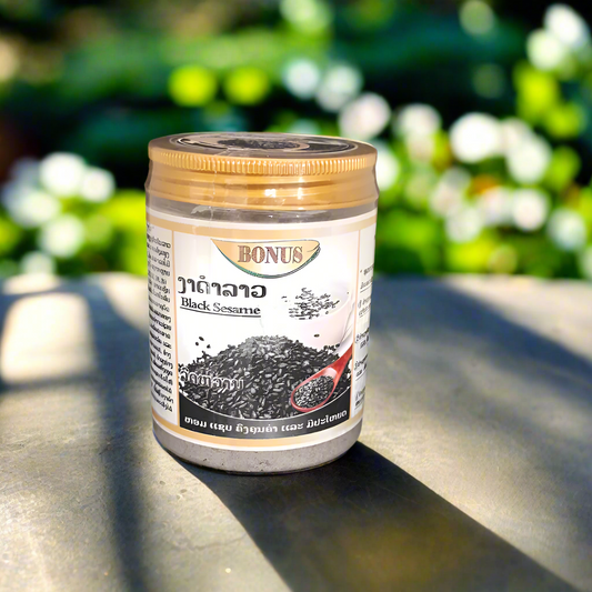 A container of Black Sesame Lao White Sesame Powder placed on a surface with a background of plants, with the label displaying the product name and an image of sesame seeds.