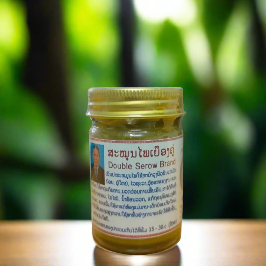 A 50g glass jar of DOUBLE SEROW herbal balm with a label featuring text and a picture, placed in a natural setting with green foliage in the background.