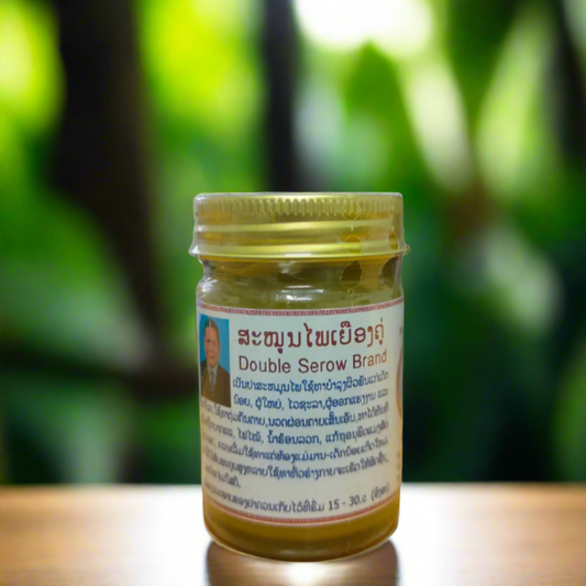 A 50g glass jar of DOUBLE SEROW herbal balm with a label featuring text and a picture, placed in a natural setting with green foliage in the background.