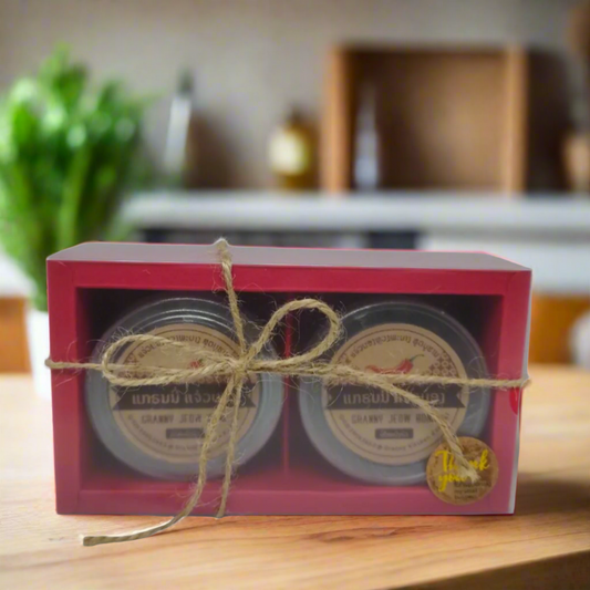 A red gift box containing two jars of Granny's Jeow Bong chili paste, tied with twine. The box is presented on a kitchen counter with a plant and various kitchen items in the background.