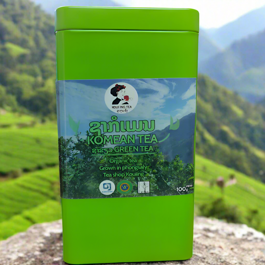 A green square metal box of Komean Organic Green Tea with a label showing the brand, product name, and an award symbol, set against a scenic background.