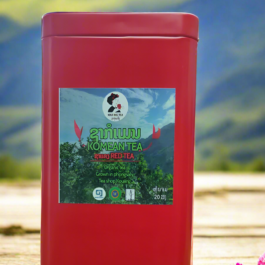 A red metal box labeled 'Komean Lao Coffee' with a graphic of tea leaves and a flower, placed on a wooden surface with a mountainous landscape in the background.