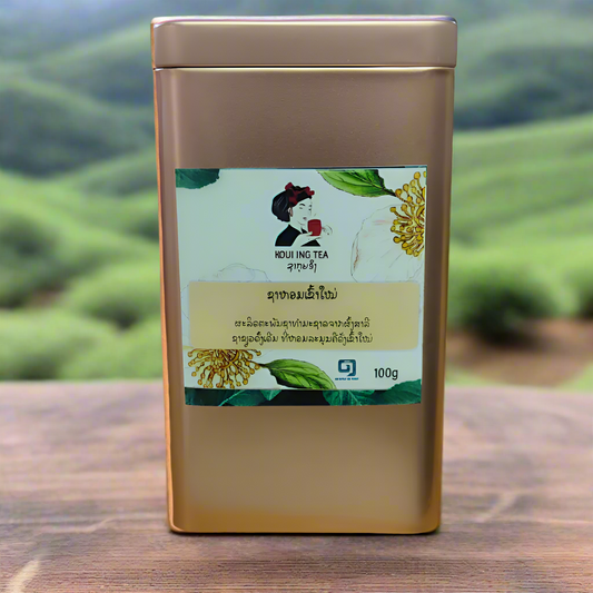 Product shot of a 100g bronze-colored metal box of Komean Koui Ing Loose Leaf Tea on a white background. The box is adorned with intricate traditional Lao patterns and text that reads "Koui Ing Tea" and "Premium Quality".