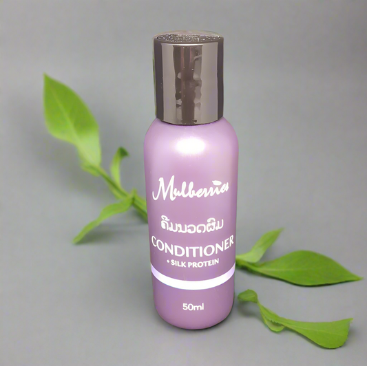 A bottle of Mulberries Silk Protein Conditioner, 50ml, with a purple label and cap, placed on a grey background with a green leaf beside it.