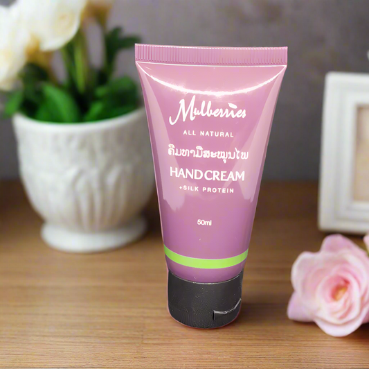 A tube of Mulberries All Natural Hand Cream with Silk Protein, placed on a wooden surface next to a vase with flowers and a framed picture.