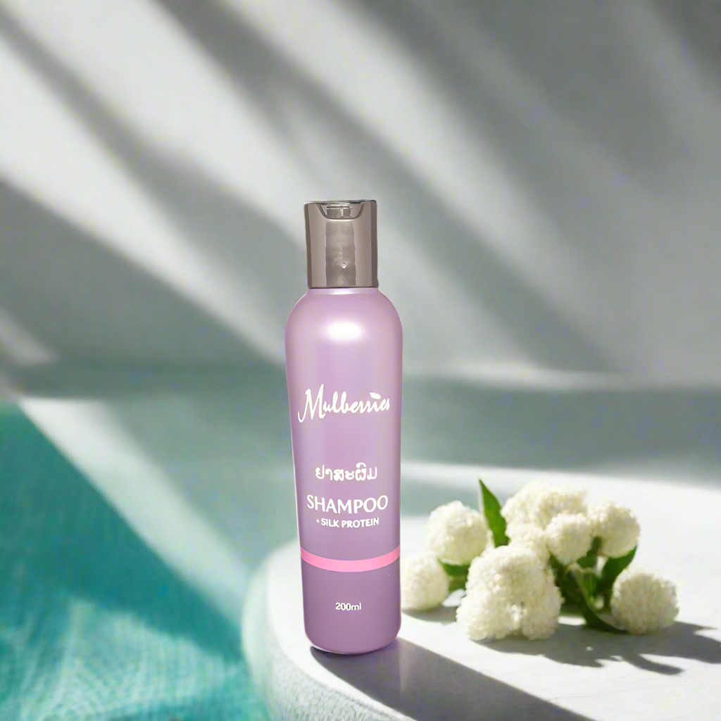 A bottle of Mulberries Silk Protein Shampoo placed on a surface with a white flower and a shadow falling over part of the image.