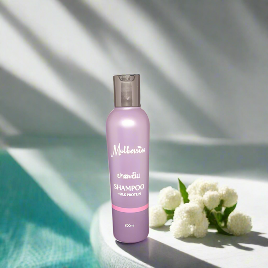 A bottle of Mulberries Silk Protein Shampoo placed on a surface with a white flower and a shadow falling over part of the image.