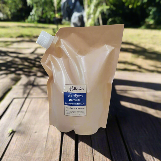 A beige sachet of Mulberries laundry detergent placed on a wooden surface with a plant background.