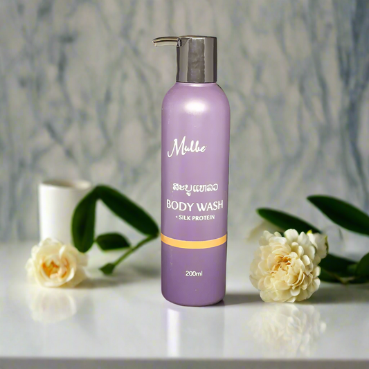 A bottle of Mulberries Body Wash with Silk Protein, 200ml, placed on a surface with a white flower and leaves in the background. The bottle is purple with a silver cap and has the brand name 'Mulie' written on it.