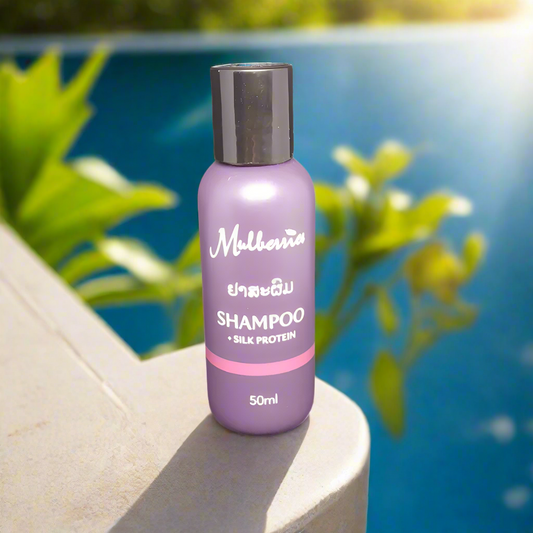 A bottle of Mulberries Shampoo with Silk Protein, 50ml, placed on a surface with a pool and plants in the background.
