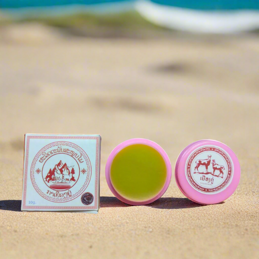 Two containers of YUANGKHU Herbal Balm lying on a sandy surface with the ocean in the background. One container is open, showing a greenish balm, while the other is closed.