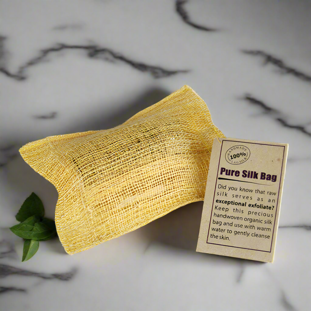 A yellow silk scrub bag with a label that reads 'PURE SILK BAG' on a marble surface, accompanied by a small green leaf.