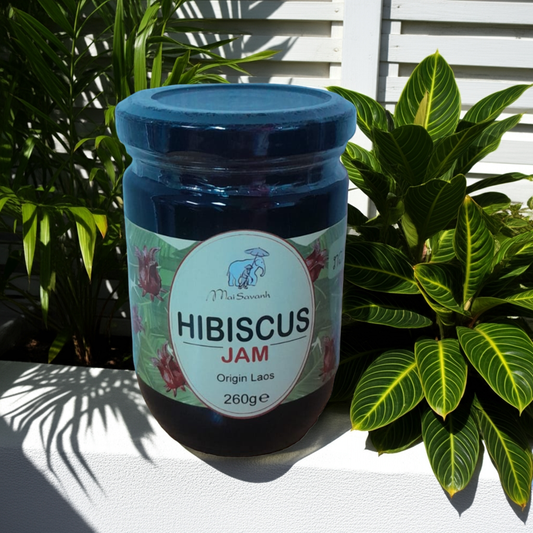 Jar of hibiscus jam with a blue lid on a white surface with plants in the background